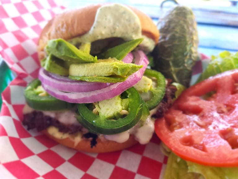 Gourmet burger with fresh lettuce, jalapeños, purple onions, and toppings on a bun, served on pink gingham paper with tomato slices and pickle