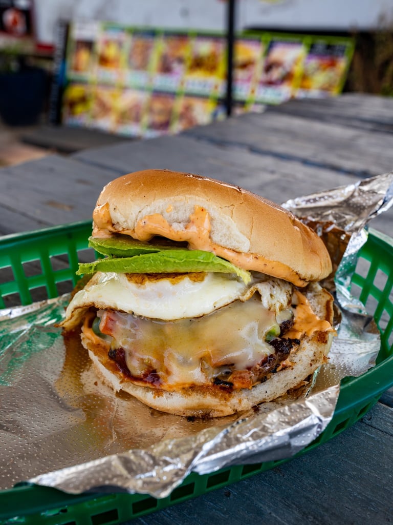 Gourmet burger with melted cheese, fried egg, avocado, and crispy patty on toasted bun served on foil-lined green basket at food stand
