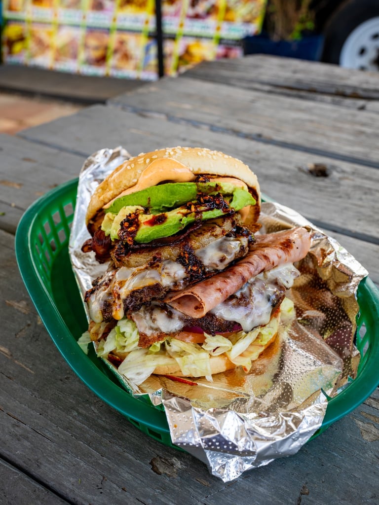 Stacked burger with avocado, bacon, and cheese on a bun, served on a green plate with fries, placed on a wooden table